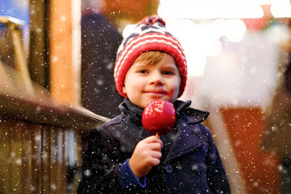 Roztomilý malý usměvavý školáček na německém vánočním trhu. Šťastné dítě v zimním oblečení jíst sladké sladké glazované jablko na pozadí se světly. Rodina, tradice, koncepce oslav — Stock fotografie
