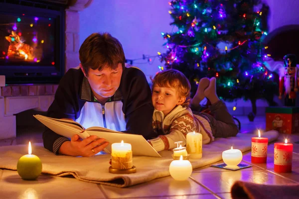 Unga far och hans lilla pojken läsa bok av en öppen spis på c — Stockfoto