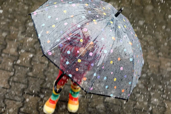 雨の日に大きな傘で遊んでいる小さな幼児の女の子。雨の中を走る幸せな正の子、水たまり。雨の服やゴムブーツを持つ就学前の子供。悪天候の日の子供の活動. — ストック写真