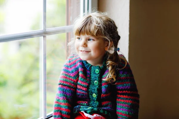 Kleines Mädchen am Fenster sitzend. Vorschulkinder tragen kuscheligen selbstgestrickten Wollpullover. Kleinkind beobachtet bei Regen draußen. Herbst und Winter. — Stockfoto