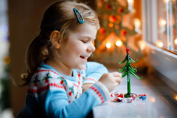 Niña pequeña sentada junto a la ventana y decorando un pequeño árbol de Navidad de vidrio con pequeños juguetes de Navidad. Feliz niño sano celebrar la fiesta tradicional familiar. Adorable bebé.. — Foto de Stock