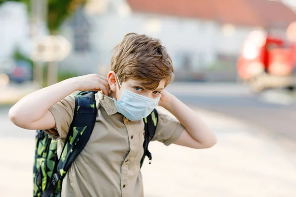 Glücklicher Junge mit Brille und medizinischer Maske wegen Coronavirus-Pandemie. Schulkind mit Schulranzen wartet am sonnigen Tag auf dem Schulweg auf den Bus. Gesundes Kind auf der Straße. — Stockfoto