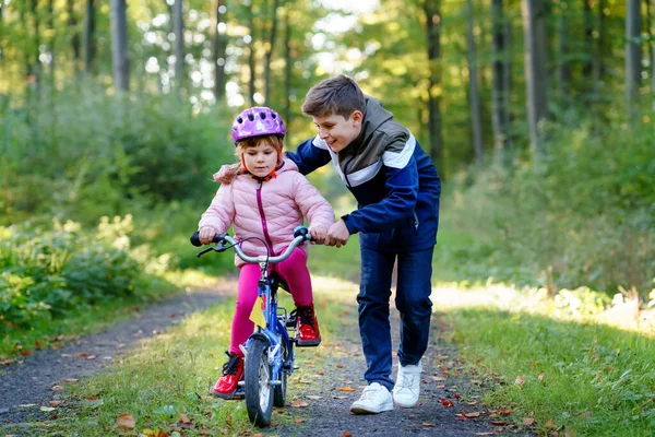 可爱的学龄前小女孩在安全头盔骑自行车 学童男孩 哥哥教快乐健康的姐姐骑自行车和玩自行车 户外运动兄弟姐妹家庭 — 图库照片