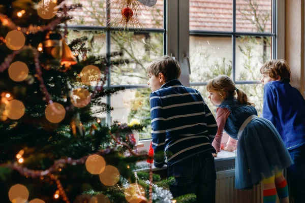 Tři roztomilé děti sedí u okna na Štědrý večer. Dva školáci a batolata, sourozenci, co vypadají venku a sní. Rodinné štěstí na tradiční dovolené — Stock fotografie