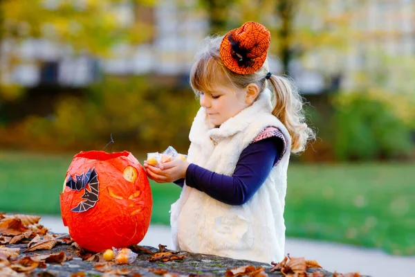 Boszorkánytrükknek öltözött, vagy halloweenkor csínynek öltözött kislány. Boldog gyermek a szabadban, narancssárga vicces kalappal és tök táskával az édes kísértéshez. Családi fesztivál szezon októberben. Szabadtéri tevékenység — Stock Fotó