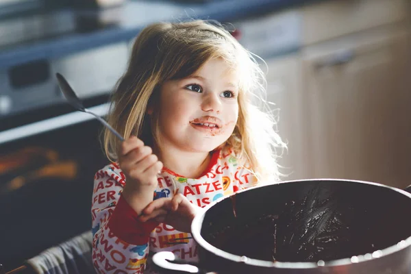 Menina da criança bonito comer massa de chocolate descansa com colher e dedos de panela. Feliz criança lambendo massa doce para muffins ou bolo, ajudando na cozinha em casa, dentro de casa. — Fotografia de Stock