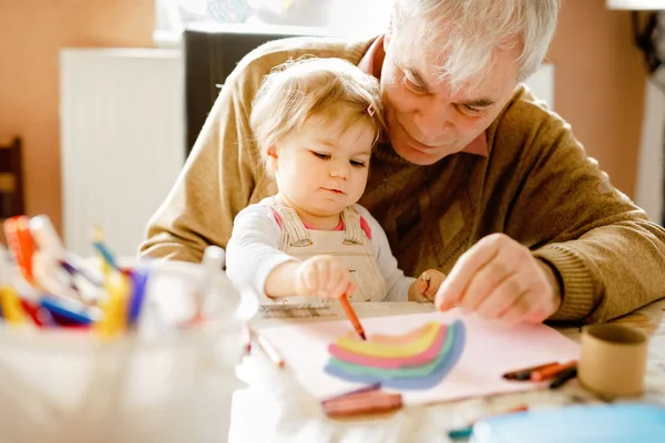 Cute little baby toddler girl and handsome senior grandfather painting with colorful pencils at home. Grandchild and man having fun together. Family and generation in love — Stock Photo, Image