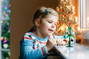 Küçük kız pencerenin kenarında oturuyor ve küçük cam Noel ağacını küçük Noel oyuncaklarıyla süslüyor. Mutlu sağlıklı çocuk geleneksel aile bayramını kutluyor. Sevimli bebek.