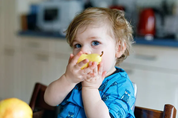 Menina adorável bonito bebê comendo pêra fresca. Criança feliz com fome de um ano segurando frutas. Menina na cozinha doméstica, tendo refeição saudável lanche. Criança loira sorrindo em casa ou no berçário — Fotografia de Stock