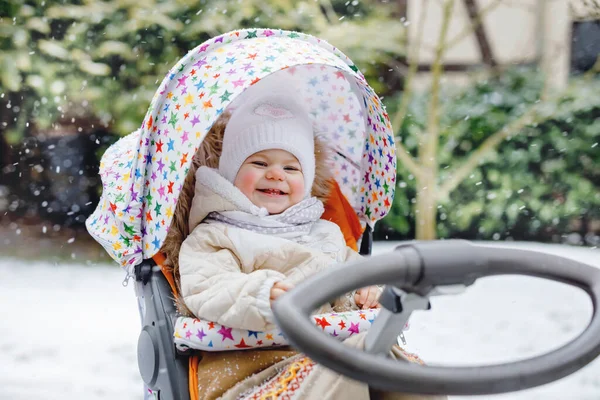 Cute little beautiful baby girl sitting in the pram or stroller on cold snowy winter day. Happy smiling child in warm clothes, fashion stylish baby coat. Babys first snow. Winter walk outdoors. — Stock Photo, Image