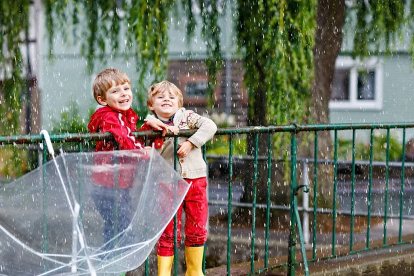 Two little boys, best friends and siblings walking with big umbrella outdoors on rainy day. Preschool children having fun. Lovely kids with rubber boots. Family outdoor leisure on bad weather day — Stock Photo, Image
