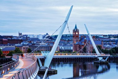 Derry, İrlanda. Kuzey İrlanda 'da, Derry Londonderry' de, Kültür Şehri 'nde şehir merkezinde aydınlatılmış bir barış köprüsü. Gece bulutlu gökyüzü, alacakaranlıkta nehirde yansıyor.