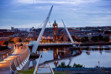 Derry, İrlanda. Kuzey İrlanda 'da, Derry Londonderry' de, Kültür Şehri 'nde şehir merkezinde aydınlatılmış bir barış köprüsü. Gece bulutlu gökyüzü, alacakaranlıkta nehirde yansıyor.