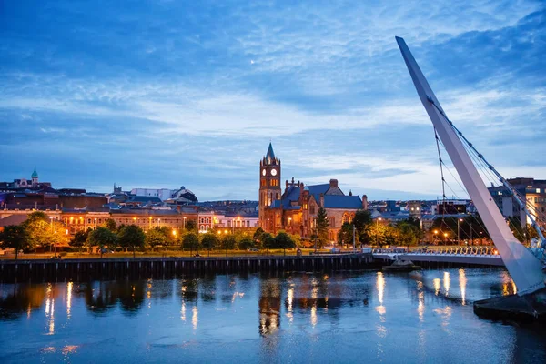 Derry, Ιρλανδία. Φωτισμένη γέφυρα ειρήνης στο Derry Londonderry, Πολιτιστική Πόλη, στη Βόρεια Ιρλανδία με το κέντρο της πόλης στο παρασκήνιο. Νύχτα συννεφιά ουρανό με αντανάκλαση στο ποτάμι στο σούρουπο — Φωτογραφία Αρχείου