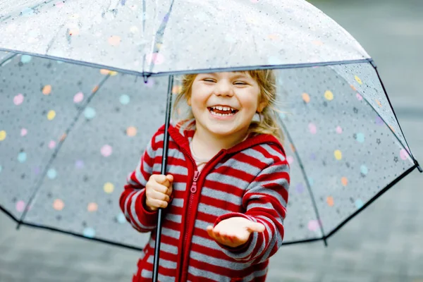 En liten småbarnstjej som leker med ett stort paraply på regnig dag. Lyckligt positivt barn som springer genom regn, pölar. Förskolebarn med regnkläder och gummistövlar. Barnaktivitet på dåligt väder dag. — Stockfoto