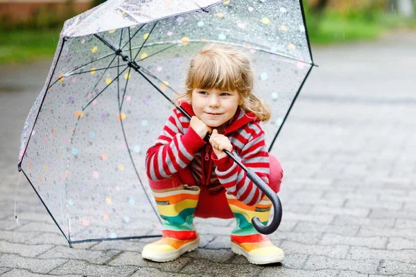 Mała dziewczynka bawiąca się dużym parasolem w deszczowe dni. Szczęśliwe pozytywne dziecko biegające w deszczu, kałuże. Dzieciak w przedszkolu z deszczowymi ubraniami i gumowymi butami. Aktywność dzieci w zły dzień pogodowy. — Zdjęcie stockowe