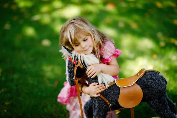 Malá školačka objímající se s houpající se koňskou hračkou. Šťastné dítě v princezně šaty na slunné letní den v zahradě. Dívka v lásce se svým oblíbeným starým veteránem hračka zvíře. — Stock fotografie
