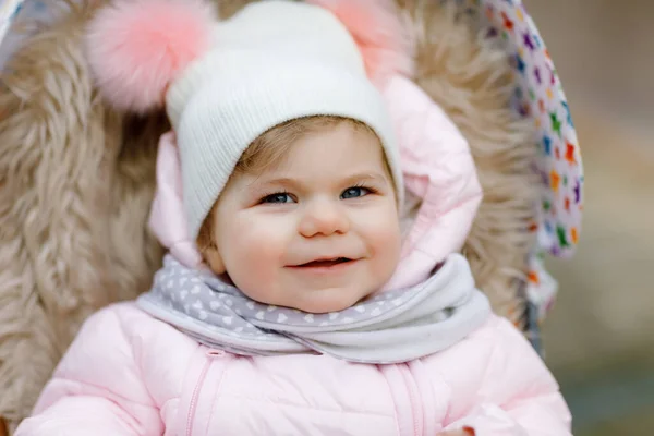 Cute little beautiful baby girl sitting in the pram or stroller on cold autumn, winter or spring day. Happy smiling child in warm clothes, fashion stylish baby coat and hat. Snow falling down — Stock Photo, Image