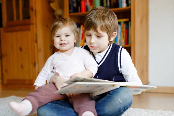 En skolegutt som leser bok for småbarnsjente, to søsken som sitter sammen og leser bøker. Vakker familie i kjærlighet, søt baby og barn som har det gøy hjemme, inne. – stockfoto