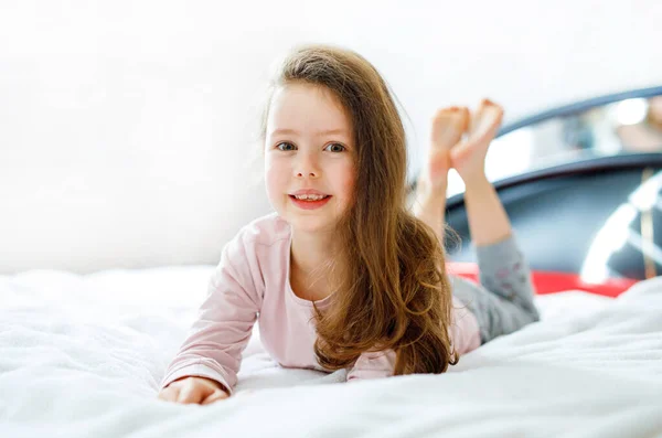 Imádnivaló, boldog kislány hosszú hajjal, miután a fehér szállodai ágyában aludt színes hálóingben.. — Stock Fotó