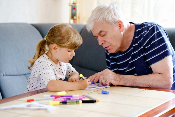 Симпатичная маленькая дошкольница и красивый дедушка, рисующий с разноцветными войлочными ручками и карандашами дома. Внук и мужчина веселятся вместе, творческая семья. Активность в помещении с ребенком. — стоковое фото