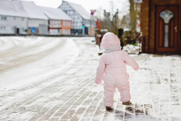 Happy little baby girl making first steps outdoors in winter through snow. Cute toddler learning walking. Child having fun on cold snowy day. Wearing warm baby pink clothes snowsuit and bobbles hat. — Stock Photo, Image