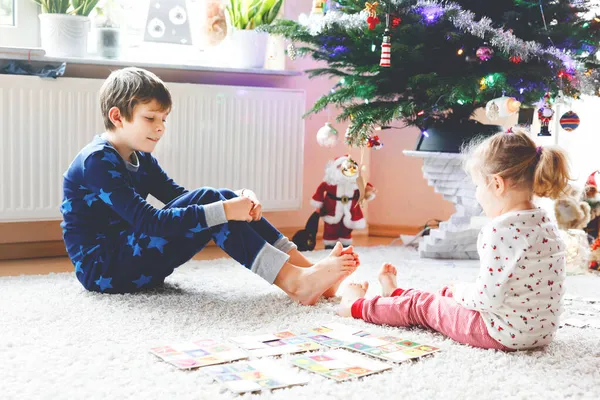 Dvě malé chilren, roztomilé batole dívka a školní kluk hrát spolu karetní hru podle zdobené vánoční stromeček. Šťastní zdraví sourozenci, bratr a sestra se spolu baví. Rodinné oslavy Vánoc — Stock fotografie