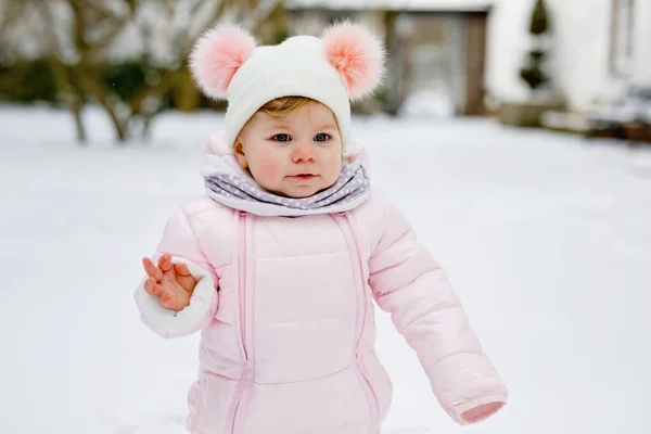 Lycklig liten flicka gör första stegen utomhus på vintern genom snö. Söta småbarn som lär sig gå. Barn har kul på kall snöig dag. Bär varma baby rosa kläder snödräkt och bubblor hatt. — Stockfoto