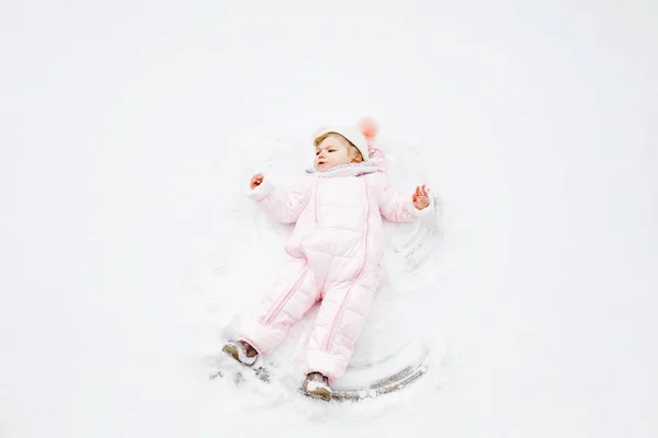Linda niña en ropa de invierno de colores haciendo ángel de nieve, acostado en la nieve. Activo ocio al aire libre con niños en invierno. Niño en traje de nieve rosa . — Foto de Stock