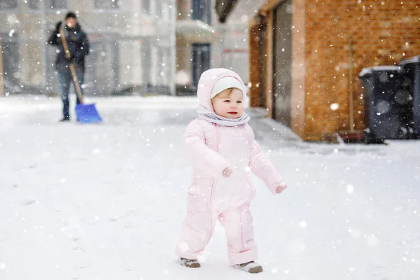 Adorable little baby girl making first steps outdoors in winter through snow. Cute toddler child learning walking. On background father, young man cleaning street with snow shovel. Happy family — Stock Photo, Image