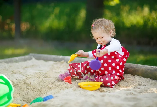 Menina bonito brincando na areia no parque infantil ao ar livre. Bebê bonito em calças de goma vermelha se divertindo no dia de verão quente ensolarado. Criança com brinquedos coloridos de areia. Bebê ativo saudável ao ar livre joga jogos — Fotografia de Stock