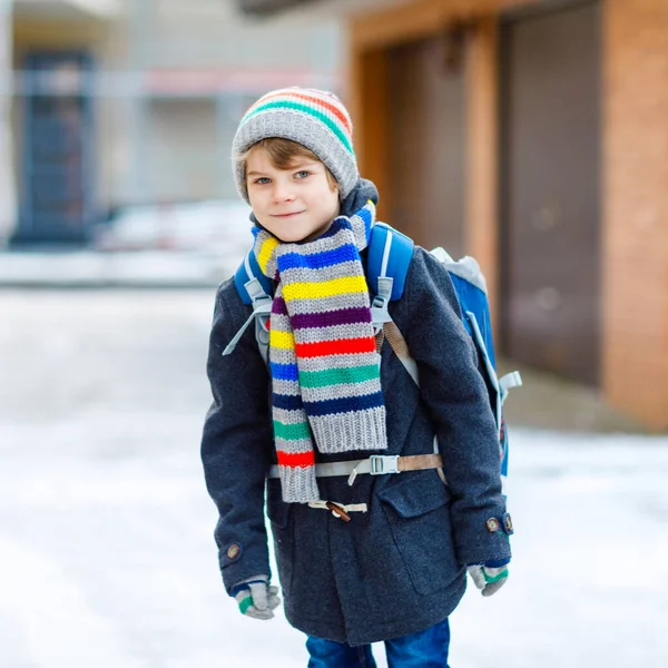 Маленький школьник начального класса, идущий в школу во время снегопада. Счастливый ребенок развлекается и играет с первым снегом. Студент с рюкзаком или сумкой в красочной зимней одежде. — стоковое фото