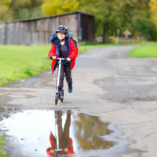 Netter kleiner Schuljunge, der auf seinem Roller auf dem Weg zur oder von der Schule fährt. Schulkind von 7 Jahren fährt durch Regenpfütze. Lustiges fröhliches Kind in bunten Kleidern und mit Helm. — Stockfoto