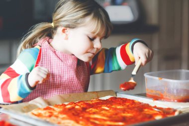 Sevimli küçük kız evde İtalyan pizzası yapıyor. Ev mutfağında, evde eğlenen sevimli mutlu çocuk. Çocuk, anaokulu öğrencisi sağlıklı yemek hazırlıyor ve yardım ediyor.