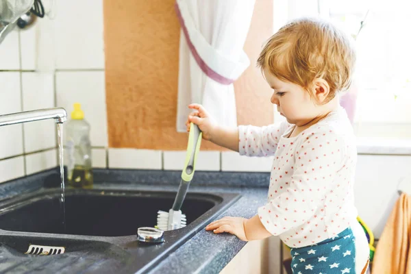 Adorable little blond baby girl washing dishes in domestic kitchen. — Stock Photo, Image