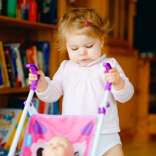 Linda menina adorável fazendo primeiros passos com carruagem de boneca. Criança bonita empurrando carrinho com brinquedo em casa. Filha feliz aprendendo andando e de pé — Fotografia de Stock