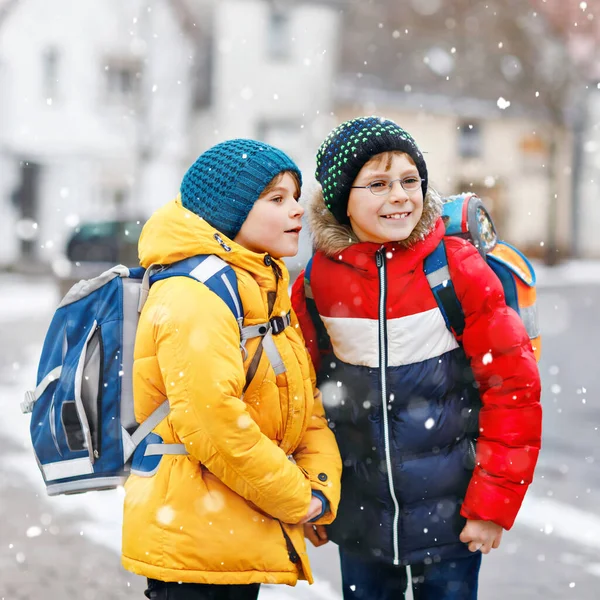 İlköğretim sınıfından iki çocuk kar yağışı sırasında okula yürüyorlar. Mutlu çocuklar eğleniyor ve ilk karla oynuyorlar. Kardeşler ve renkli kış kıyafetleri içinde sırt çantalı en iyi arkadaşlar.. — Stok fotoğraf