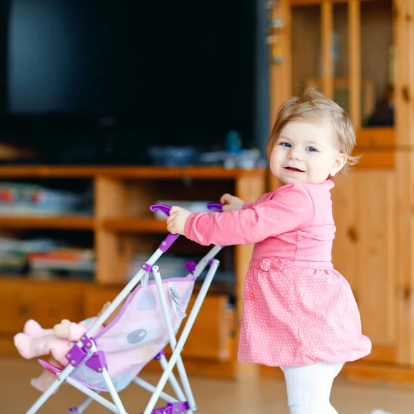 Schattig schattig baby meisje maken eerste stappen en duwen met pop rijtuig. Mooi peuter kind duwen kinderwagen met speelgoed thuis. Gelukkig kind met twee poppen — Stockfoto