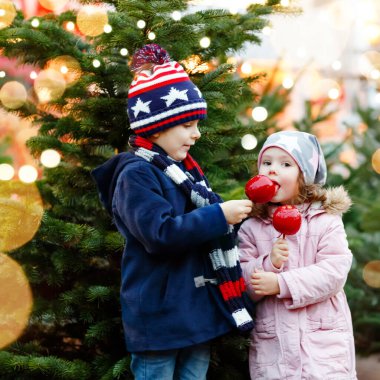 İki küçük gülümseyen çocuk, anaokulu çocuğu ve kız Alman Noel pazarında şekerli elma yiyorlar. Arka planda ışıkları ve xmas ağaçları olan mutlu kardeşler. Ailenin komik kardeşleri..