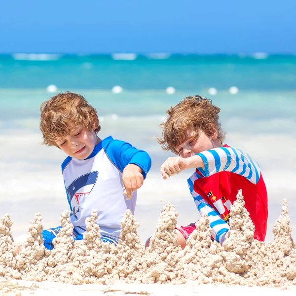 Dos niños divirtiéndose con la construcción de un castillo de arena en la playa tropical en la isla. Niños sanos jugando juntos en sus vacaciones. Gemelos, hermanos felices riendo y sonriendo —  Fotos de Stock