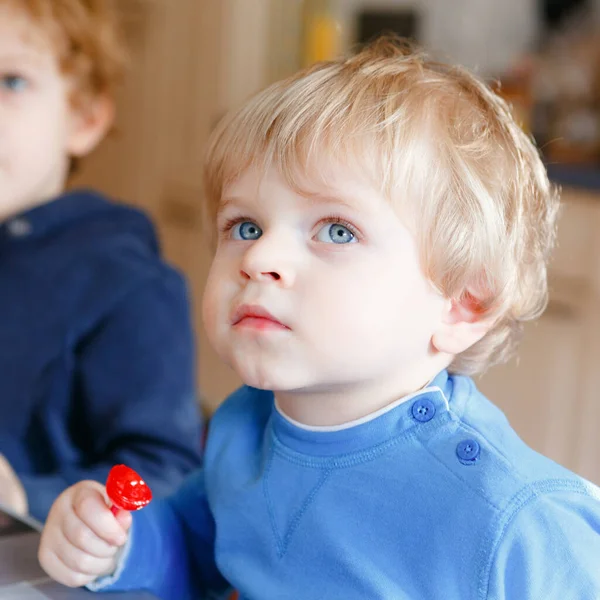 Due bambini piccoli, bambini fratelli carini che guardano cartoni animati in TV e mangiano caramelle lolipop. Fratelli felici insieme all'asilo o all'asilo nido, a pranzo. — Foto Stock