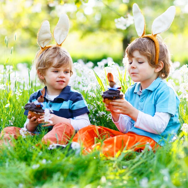Два маленьких мальчика в пасхальных кроликах едят шоколадные торты и кексы — стоковое фото