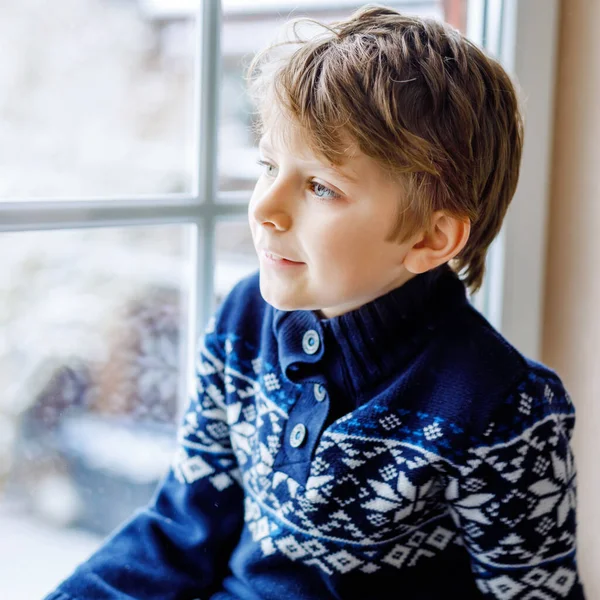 Niño adorable feliz sentado cerca de la ventana y mirando hacia fuera en la nieve en el día de Navidad o la mañana. Sonriente niño sano fascinado observando nevadas y grandes copos de nieve — Foto de Stock