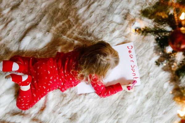 Noel ağacının altında pijamalarıyla küçük şirin bir anaokulu kızı Noel Baba 'ya evde, evde dilek listesi mektupları yazıyor. Geleneksel Hıristiyan festivali. Mutlu bebek Noel 'de hediye bekliyor.. — Stok fotoğraf