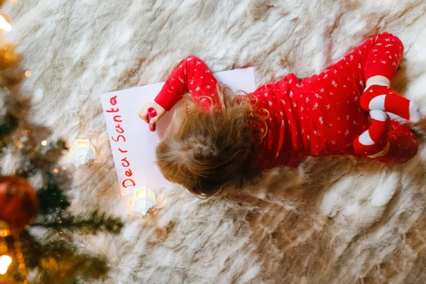 Μικρό χαριτωμένο κορίτσι προσχολικής ηλικίας με πιτζάμες κάτω από το χριστουγεννιάτικο δέντρο, γράφοντας επιστολή λίστα επιθυμιών στον Άγιο Βασίλη στο σπίτι, σε εσωτερικούς χώρους. Παραδοσιακό Χριστιανικό φεστιβάλ. Ευτυχισμένο μικρό παιδί περιμένει δώρα για τα Χριστούγεννα. — Φωτογραφία Αρχείου