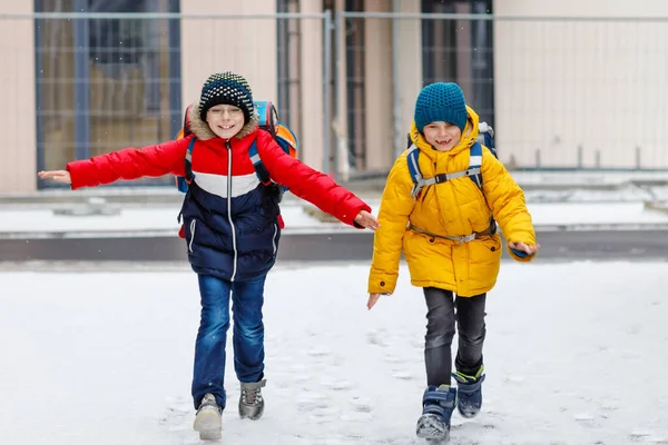 Två små pojkar i grundskolan som går till skolan under snöfallet. Glada barn som har roligt och leker med första snön. Syskon och bästa vän med ryggsäck i färgglada vinterkläder. — Stockfoto