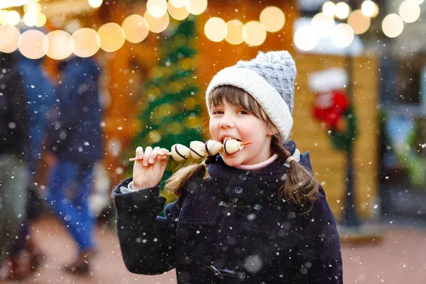 Little cute kid girl eating white chocolate covered strawberries on skewer on traditional German Christmas market. Happy child on traditional family market in Germany during snowy day. — Stock Photo, Image