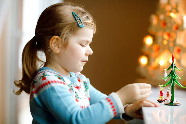 Niña pequeña sentada junto a la ventana y decorando un pequeño árbol de Navidad de vidrio con pequeños juguetes de Navidad. Feliz niño sano celebrar la fiesta tradicional familiar. Adorable bebé.. — Foto de Stock