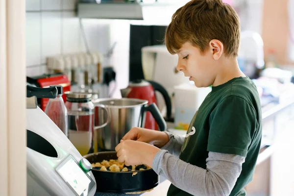 Menino da escola loira feliz cozinhando bolo de ameixa na cozinha doméstica, dentro de casa. Engraçado linda criança pré-adolescente saudável se divertindo com a ajuda. — Fotografia de Stock