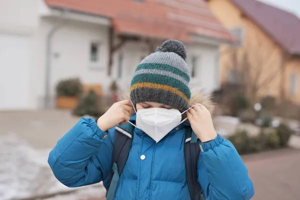 Rapaz a usar máscara médica a caminho da escola. Mochila infantil mochila. Schoolkid no outono frio ou dia de inverno com roupas quentes. Fechamento e tempo de quarentena durante a doença pandémica da coroa — Fotografia de Stock